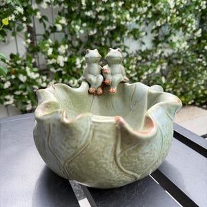 May include: A ceramic bowl in the shape of a leaf, with two small frog figurines perched on the edge. The bowl is a muted green color with a textured surface. The frogs have light green bodies and orange feet.