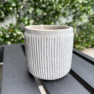 May include: A small, light gray, textured ceramic planter with vertical grooves. The round pot sits on a dark gray surface, with a blurred background of green foliage and white flowers.