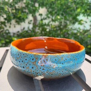 May include: A handmade ceramic bowl with a vibrant design. The bowl features a speckled turquoise exterior and a glossy orange interior. The bowl is round with a slightly irregular shape, perfect for serving or display.