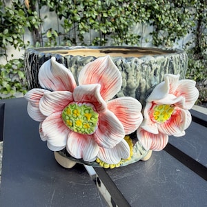 May include: A ceramic planter with a textured, dark green exterior featuring two large, three-dimensional lotus flowers. The flowers have white petals with pink edges and bright yellow and green centers. The planter stands on small feet.