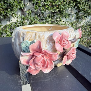 May include: A ceramic planter with a textured, cream-colored exterior resembling tree bark. The vessel is adorned with three-dimensional, glossy pink ceramic roses and green leaves attached to the side, featuring a smooth, light-colored interior.