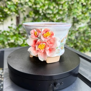 May include: A ceramic flower pot with a light purple rim and a floral design. The pot features three-dimensional pink flowers with yellow centers. The pot is resting on a black rotating platform.