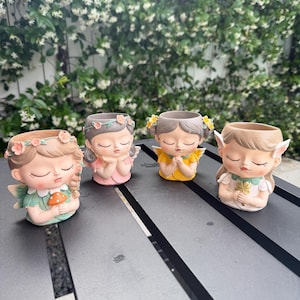 May include: Four small, decorative fairy planters with closed eyes. Each planter features a different design, including flower crowns, wings, and colorful clothing. The planters are arranged on a dark gray surface, with a backdrop of green foliage and white flowers.