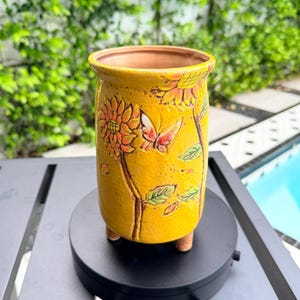 May include: Yellow ceramic planter with a sunflower and butterfly design. The planter has a cylindrical shape, three small feet, and a textured surface. The sunflower and butterfly are in relief, with orange and brown accents. Ideal for small plants.