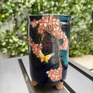 May include: A dark blue ceramic planter with a hand-painted cherry blossom design. The planter features a brown tree trunk, pink flowers, and a yellow butterfly. It stands on three small wooden feet.