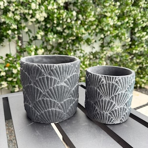May include: Two dark gray, textured ceramic planters with a fan-like pattern. The planters are different sizes and sit on a dark gray table. The background features a wall covered in green foliage and white flowers.