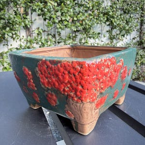 May include: A square ceramic planter featuring a dark green base glaze with a prominent, textured bright red drip pattern on the exterior. The vessel is supported by four short, sturdy feet and has a smooth, light brown interior finish.