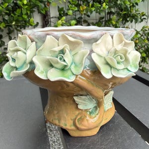 May include: A ceramic planter featuring three dimensional sculpted pale green roses around the rim. The base is a textured brown finish with sculpted green leaves. The interior of the bowl has a glossy, iridescent finish with soft blue and white tones.