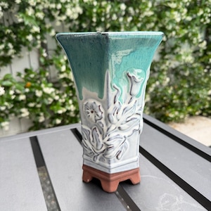 May include: A square ceramic vase with a teal and white glaze. The vase features a floral relief design and a brown base. The vase is sitting on a gray table.