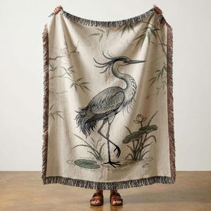 May include: Beige throw blanket featuring a detailed illustration of a heron standing in a pond with aquatic plants. The blanket has a fringed edge in shades of brown and beige. The heron is depicted in shades of gray and blue.