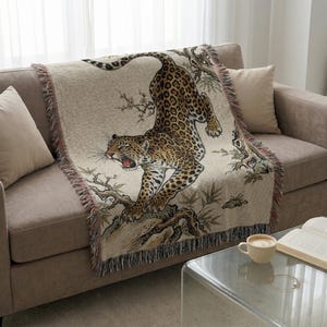 Pode incluir: Um cobertor bege com uma ilustração detalhada de um leopardo com pelagem manchada, contra um fundo de galhos e folhagens. O cobertor tem uma borda com franjas e está sobre um sofá.