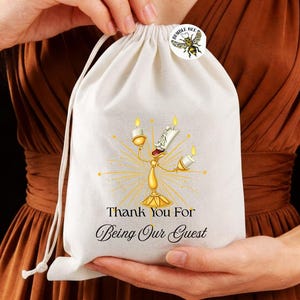 May include: A white drawstring bag featuring a gold candelabra illustration with the text "Thank You For Being Our Guest". A small round sticker with a bumble bee graphic is attached to the bag.