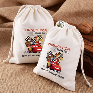 K&ouml;nnte beinhalten: Zwei cremefarbene Kordelzugbeutel mit einem rot-schwarzen Rennwagendesign mit Zeichentrickfiguren. Die Beutel tragen den Text "THANKS FOR Racing By..." und "TONY 5TH BIRTHDAY PARTY!". Die Beutel befinden sich auf einer braunen Oberfl&auml;che.