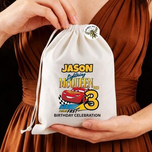 Puede incluir: Una bolsa blanca con cord&oacute;n, con un dise&ntilde;o colorido de Rayo McQueen, el nombre "Jason" y el n&uacute;mero "3". El texto "Think Fast Birthday Celebration" tambi&eacute;n est&aacute; impreso. Una peque&ntilde;a pegatina redonda con una abeja est&aacute; adherida en la parte superior.