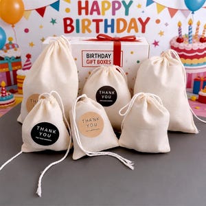 K&ouml;nnte beinhalten: Eine Sammlung beiger Kordelzugbeutel mit wei&szlig;en Kordeln, einige mit "THANK YOU" Aufklebern. Im Hintergrund eine wei&szlig;e Box mit rotem Band und dem Text "BIRTHDAY GIFT BOXES", zusammen mit einer Geburtstagstorte und den Worten "HAPPY BIRTHDAY".
