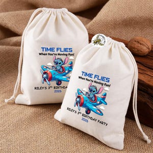 May include: Two off-white drawstring bags with a blue and red airplane graphic featuring a cartoon character. Text on the bags reads "TIME FLIES When You're Having Fun!" and "RILEY'S 3RD BIRTHDAY PARTY 2026".