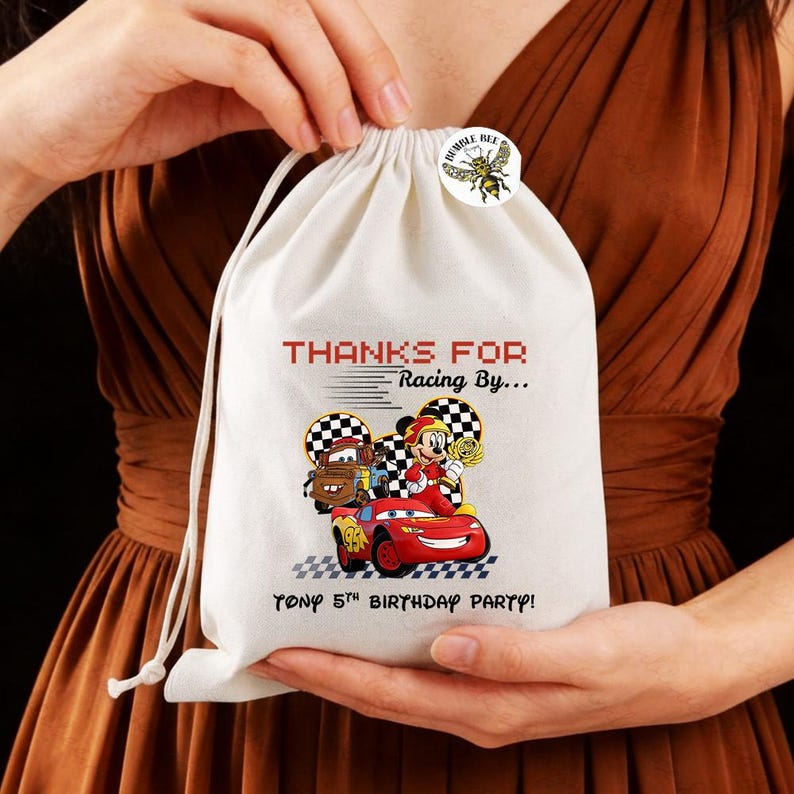 K&ouml;nnte beinhalten: Ein wei&szlig;er Kordelzugbeutel mit einer farbenfrohen Grafik von Zeichentrickautos und -figuren, mit dem Text "THANKS FOR Racing By..." und "TONY 5th BIRTHDAY PARTY!". Ein kleiner runder Aufkleber mit einer Biene ist oben angebracht.