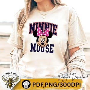 May include: Beige t-shirt featuring a Minnie Mouse graphic. The design includes the text "MINNIE MOUSE" in a navy blue and red arched font, with Minnie's face and a pink polka dot bow. The shirt has short sleeves.