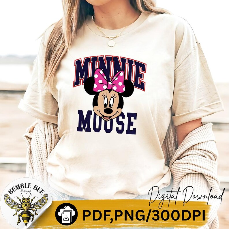 May include: Beige t-shirt featuring a Minnie Mouse graphic. The design includes the text "MINNIE MOUSE" in a navy blue and red arched font, with Minnie's face and a pink polka dot bow. The shirt has short sleeves.