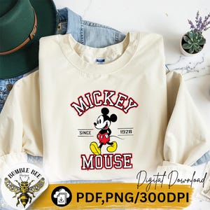 May include: Cream-colored sweatshirt featuring a Mickey Mouse graphic. The design includes the word "Mickey" in red and white, the character illustration, and the word "Mouse". The text "Since 1928" is also included.