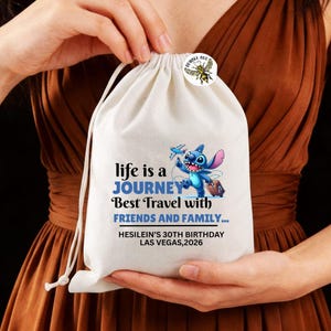 Może przedstawiać: Biała torba ze sznurkiem z napisem "life is a JOURNEY Best Travel with FRIENDS AND FAMILY..." i ilustracją postaci z kresk&oacute;wki. Do torby przypięta jest mała przypinka z grafiką pszczoły. Torba jest trzymana przez osobę.
