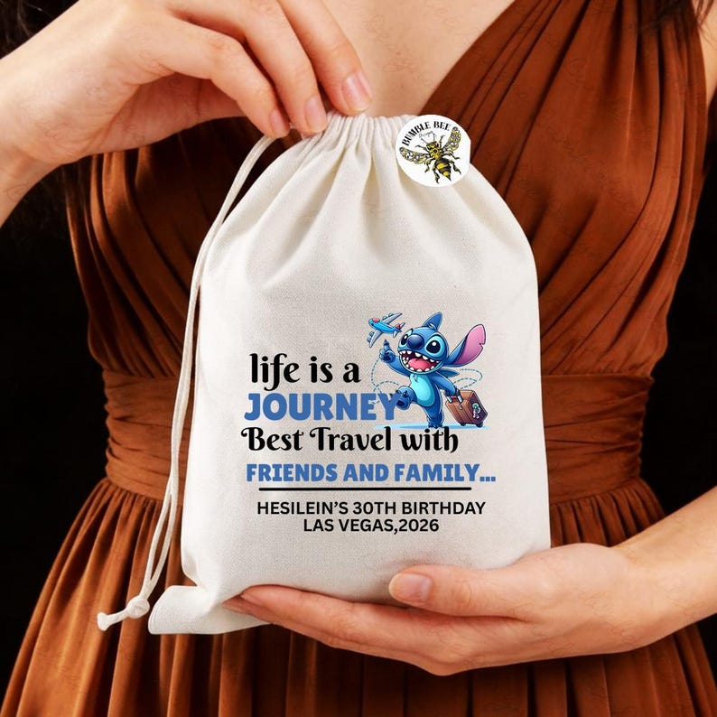 Może przedstawiać: Biała torba ze sznurkiem z napisem "life is a JOURNEY Best Travel with FRIENDS AND FAMILY..." i ilustracją postaci z kresk&oacute;wki. Do torby przypięta jest mała przypinka z grafiką pszczoły. Torba jest trzymana przez osobę.