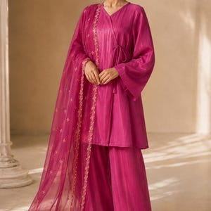 Peut inclure: Ensemble magenta en trois pièces comprenant un haut cache-cœur à manches évasées, un pantalon large et une écharpe transparente. L'écharpe est ornée de broderies dorées. L'ensemble est en tissu brillant.
