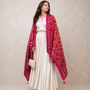 Könnte beinhalten: Eine Frau trägt ein rotes Tuch mit floralem Muster und einem leuchtend pinkfarbenen Rand. Sie trägt ein weißes Outfit und eine cremefarbene Handtasche mit goldener Kette. Das Outfit ist für besondere Anlässe und Veranstaltungen geeignet.