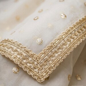May include: Close-up of a cream-colored fabric with gold embellishments. The fabric features a detailed gold border with sequins and beadwork. Scattered gold floral accents add to the elegant design. The overall aesthetic is luxurious and ornate.