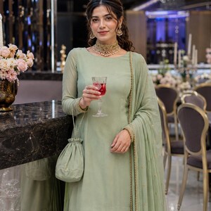 May include: A sage green long-sleeved tunic and matching pleated skirt set with a coordinating sheer dupatta featuring gold embroidery. The outfit includes a matching drawstring pouch bag, a gold choker necklace, and gold earrings with pearl accents.