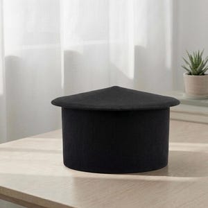Pode incluir: Um recipiente cilíndrico preto com uma tampa plana e angulada. O recipiente está sobre uma superfície de madeira clara. Uma pequena planta em vaso é visível no fundo. O design é minimalista e moderno.