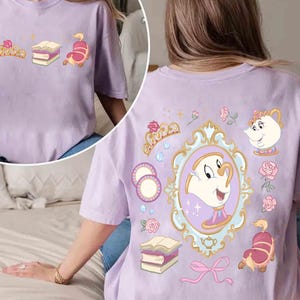 Puede incluir: Una camiseta de manga corta de color morado claro con un estampado gráfico de un personaje de taza de té dentro de un marco ornamentado, rodeado de ilustraciones de una tetera, libros, una corona, rosas y un lazo rosa en la espalda.