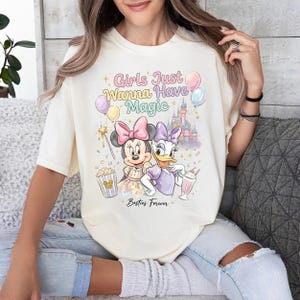 May include: Cream-colored t-shirt featuring a colorful graphic of Minnie Mouse and Daisy Duck with the text "Girls Just Wanna Have Magic" and "Besties Forever". The design includes balloons, a castle, and popcorn.