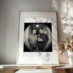 Peut inclure: Une photographie encadrée en noir et blanc présentant une vue au fisheye de deux personnes dans un couloir. L'une porte une veste claire, tandis que l'autre porte un manteau sombre et tient une bouteille. L'image est placée dans un cadre simple.
