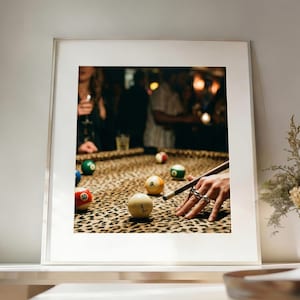 May include: A framed photograph of a pool table with a leopard print felt. Several numbered billiard balls are scattered across the table. A hand with multiple rings is poised to strike a ball with a cue stick.