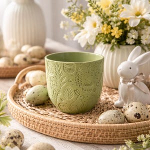 Puede incluir: Una maceta de cerámica verde con diseños en relieve con motivos de Pascua, incluyendo conejos y huevos, se encuentra sobre una bandeja tejida con huevos moteados. Jarrones blancos con flores y un conejo de cerámica completan la decoración primaveral.