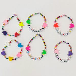 May include: Six circular bracelets made of small multicolored seed beads. The bracelets feature various plastic beads shaped like mouse heads in bright colors, and some include white letter beads spelling out the words WONDER or ALASKA.