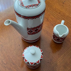 May include: A white ceramic tea set with a red and black geometric pattern. The set includes a teapot, a creamer, and a sugar bowl with a lid. The teapot has a curved spout and handle. The sugar bowl has a small white knob on the lid.