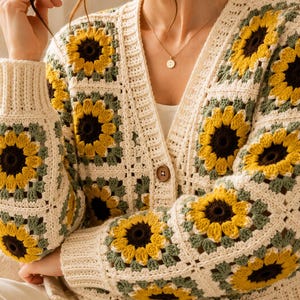 Puede incluir: Un cárdigan de ganchillo de manga larga con un patrón repetitivo de cuadrados de la abuela con motivos de girasoles en amarillo, marrón oscuro y verde salvia sobre un fondo blanco roto, con un botón marrón en el cierre frontal.