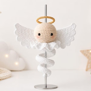 Puede incluir: Una figura de ángel tejida a ganchillo con cabeza redonda beige, ojos negros, mejillas rosadas, halo dorado y alas blancas. El ángel está montado sobre un soporte de metal plateado con una base en espiral tejida en blanco alrededor del poste.