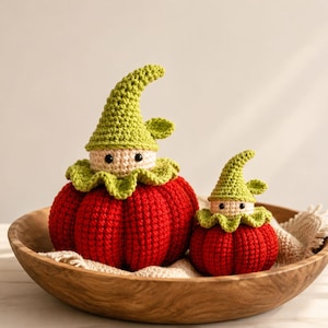 May include: Two crocheted figures shaped like pumpkins with red ribbed bodies and light green pointed hats. Each figure has a beige face with two small black bead eyes and a ruffled green collar around the neck, resting inside a wooden bowl.