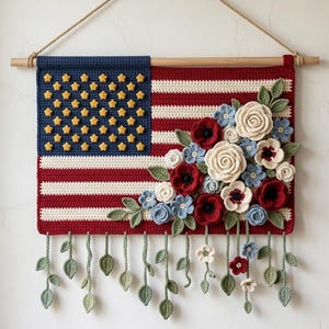 May include: A rectangular crochet wall hanging featuring the American flag pattern with red and white stripes and a blue canton adorned with yellow stars. A cluster of three-dimensional crochet flowers in red, white, and blue hangs from the side.