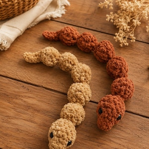 May include: Two crocheted snake toys made of soft yarn. One snake is light beige and the other is a darker rust-orange color. Both snakes feature a segmented, bumpy body texture and small, round, black plastic eyes positioned on their heads.