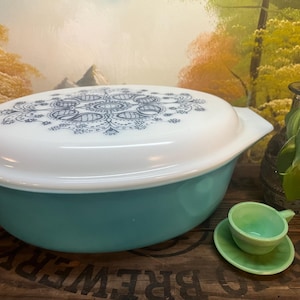 May include: A vintage turquoise and white Pyrex casserole dish with a decorative lid. The lid features a blue and white floral design. A small green teacup and saucer are in the foreground.