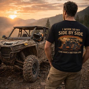 Könnte beinhalten: Schwarzes T-Shirt mit dem Text "I WORK SO MY SIDE BY SIDE CAN HAVE A BETTER LIFE" und einer Grafik eines Geländewagens. Ein schlammiger Geländewagen steht vor einer Person. Der Hintergrund zeigt einen Sonnenuntergang über den Bergen.