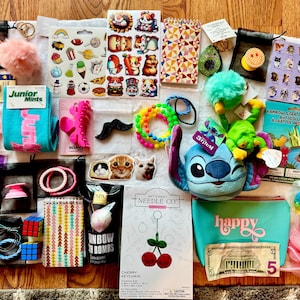 May include: A collection of various small items, including a Stitch plush, stickers, hair accessories, a Rubik's cube, a cherry keychain, and a small turquoise cosmetic bag with the word "happy".