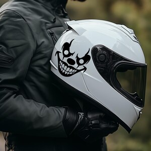 Op de afbeelding: Witte motorhelm met een zwarte clowngezichtsticker. De helm heeft een donker vizier en wordt vastgehouden door een persoon die een zwarte jas en handschoenen draagt. De achtergrond is wazig met groene bomen.