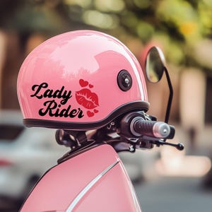 Peut inclure: Casque de moto rose avec l'inscription "Lady Rider" en noir, accompagné d'un dessin de lèvres rouges et de coeurs. Le casque est monté sur un scooter rose, avec un guidon et un rétroviseur noirs.
