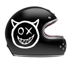 Op de afbeelding: Glanzende zwarte motorhelm met een witte duivelsgezicht-afbeelding. De afbeelding toont hoorns, een "OX"-symbool en een brede, grijnzende mond. De helm heeft een zilveren rand en een roze binnenkant.