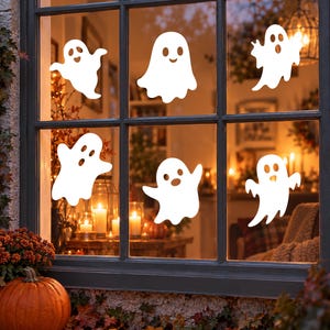 Könnte beinhalten: Ein Set aus sechs weißen, geisterförmigen Fensteraufklebern, die auf einer Glasscheibe angebracht sind. Jeder Geist hat eine einzigartige Silhouette mit ausgeschnittenen Augen und Mündern. Die Aufkleber sind blickdicht weiß und auf den Scheiben verteilt.